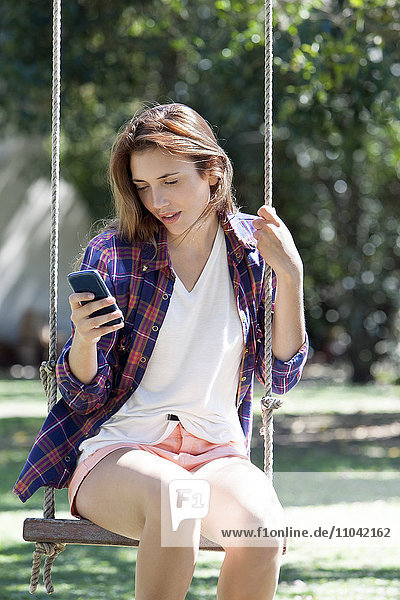 Junge Frau sitzt auf der Schaukel und schaut auf das Smartphone.