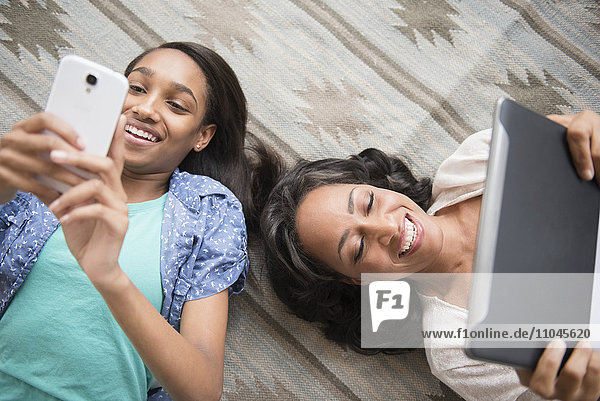 Mutter und Tochter liegen auf einem Teppich und benutzen ein Handy und ein digitales Tablet