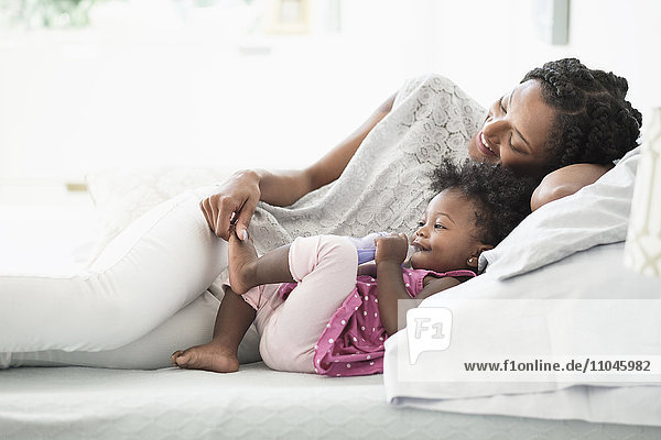 Schwarze Frau spielt mit dem Fuß ihrer kleinen Tochter auf dem Bett