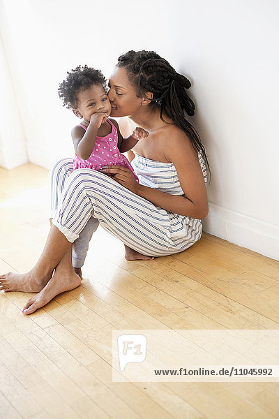 Schwarze Frau sitzt auf dem Boden und küsst die Wange ihrer kleinen Tochter