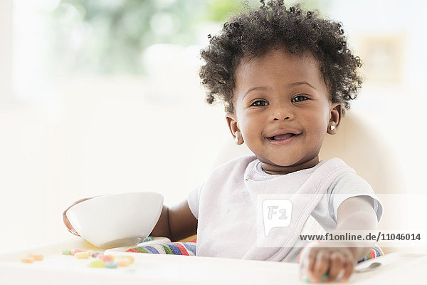 Lächelndes schwarzes Babymädchen isst Müsli aus einer Schüssel im Hochstuhl