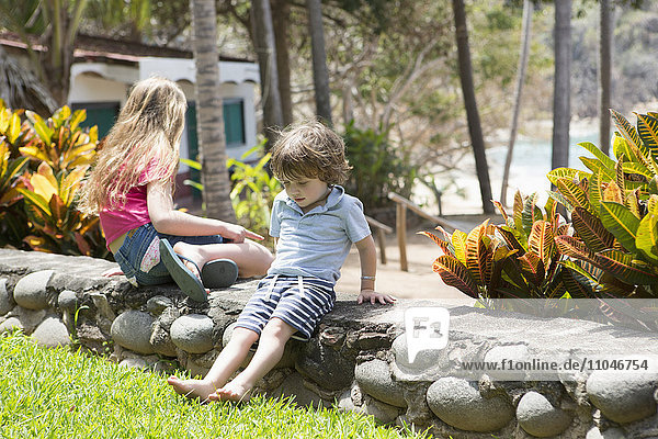 Kaukasischer Bruder und Schwester sitzen auf einer Steinmauer am Strand