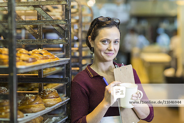Caucasian woman holding clipboard drinking coffee in bakery