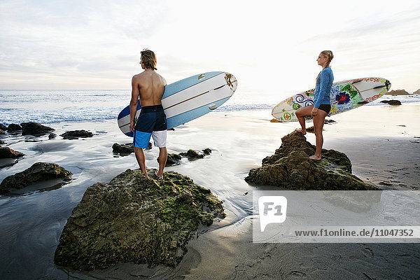 Kaukasisches Paar steht auf Felsen und trägt Surfbretter am Strand