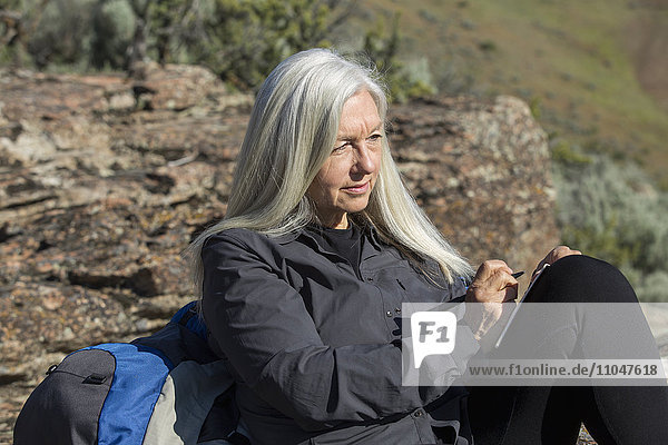 Ältere kaukasische Frau lehnt an einem Felsen und schreibt in ein Tagebuch
