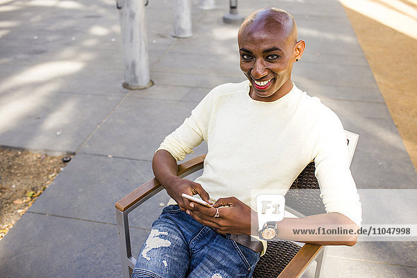 Lächelnder schwuler schwarzer Mann  der auf einem Stuhl im Freien eine SMS schreibt