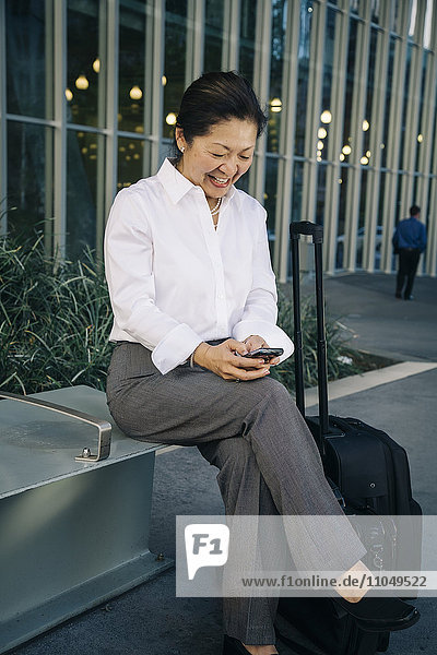 Businesswoman sitting on bench with suitcase texting on cell phone