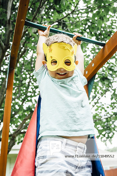 Caucasian boy wearing superhero costume hanging on monkey bars