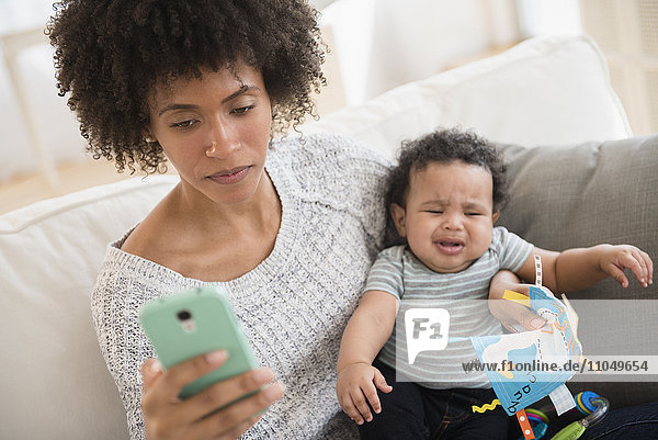 Mutter hält weinendes Baby im Arm  während sie eine SMS auf dem Handy schreibt