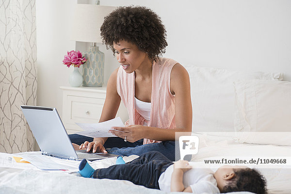 Mother paying bills on laptop with baby son on bed