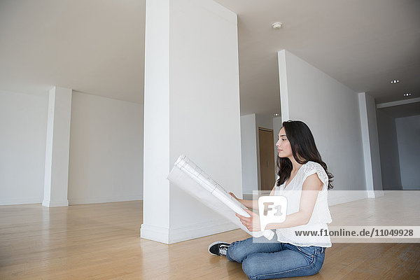 Hispanische Frau sitzt auf dem Boden einer leeren Wohnung und hält einen Bauplan