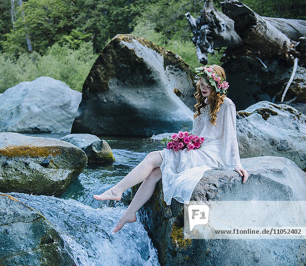 Kaukasische Frau mit Blumenkrone auf einem Felsen an einem Wasserfall