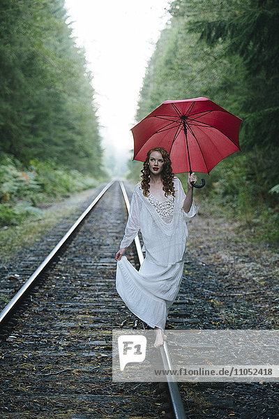 Kaukasische Frau geht mit Regenschirm im Wald auf Bahngleisen