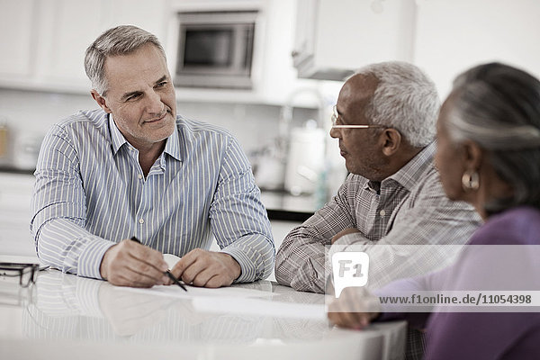Drei Personen an einem Tisch  ein älteres Ehepaar sitzt mit einem Mann zusammen  der ihnen mit Papier und Stift Informationen gibt.