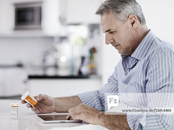 A man seated at a table using a digital tablet  holding a medicine pill bottle  and reading the label.