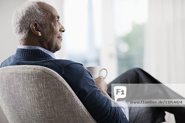 Ein reifer Mann mit grauem Haar  der sich auf einem Stuhl zurücklehnt und sich bei einer Tasse Tee oder Kaffee entspannt.