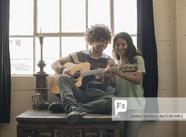 Loft-Wohnen. Ein junger Mann spielt Gitarre und neben ihm eine Frau  die mit einem Smartphone fotografiert.