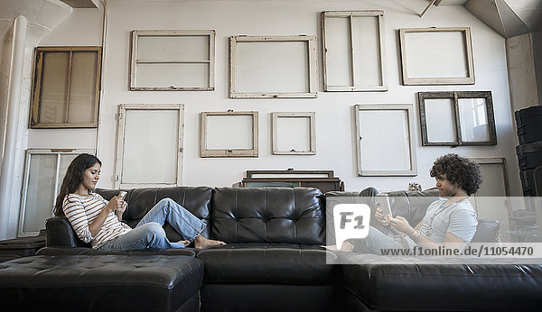Loft-Dekor. Eine Wand  an der Bilder in Rahmen aufgehängt sind  die umgekehrt die Rückseiten zeigen. Ein junges Paar sitzt auf dem Sofa  einer benutzt ein Smartphone und einer hält ein digitales Tablet.