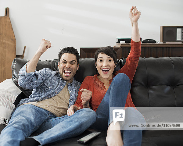 A man and woman sitting on a sofa  celebrating and pumping the air with their fists. Watching sport on the tv.