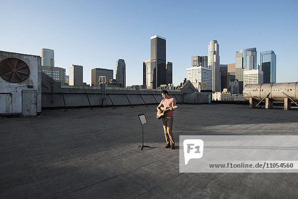 A man standing on a rooftop overlooking the city playing a guitar.