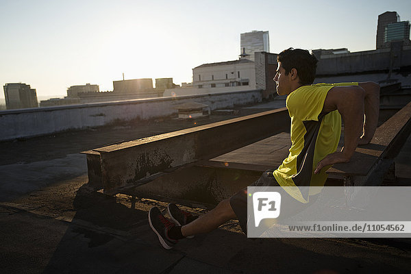 A man in exercise clothes on a rooftop overlooking the city  doing bench shoulder push ups.