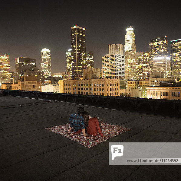 A couple sitting on a rug on a rooftop overlooking a city lit up at night.