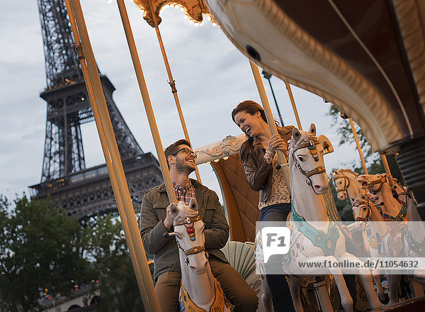 Ein Paar  Mann und Frau  reiten auf traditionellen Galoppern auf einem Karussell im Schatten des Eiffelturms in Paris.