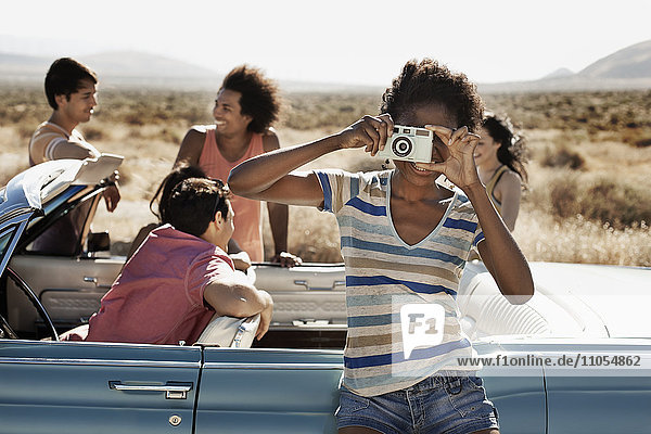 A group of friends by a pale blue convertible on the open road  on a flat plain surrounded by mountains  one holding a camera.