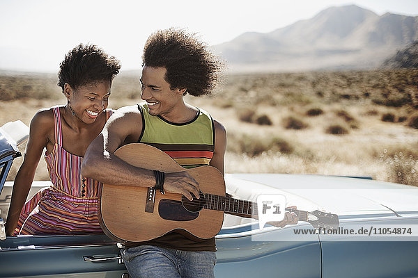 A young couple standing by a pale blue convertible on the open road  the man playing a guitar.