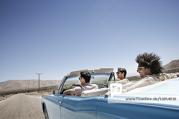 Three young people in a pale blue convertible car  driving on the open road across a flat dry plain