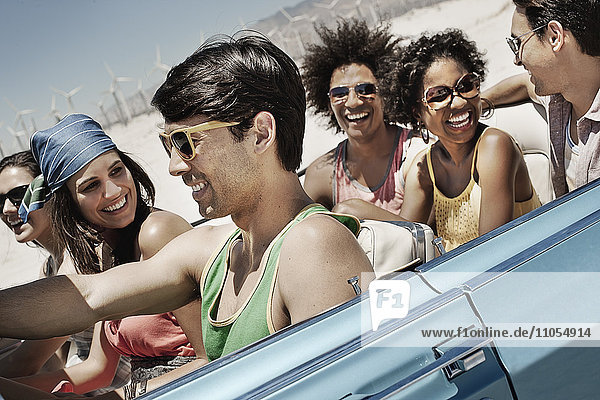 A group of friends in a pale blue convertible on the open road  driving across a dry flat plain surrounded by mountains.
