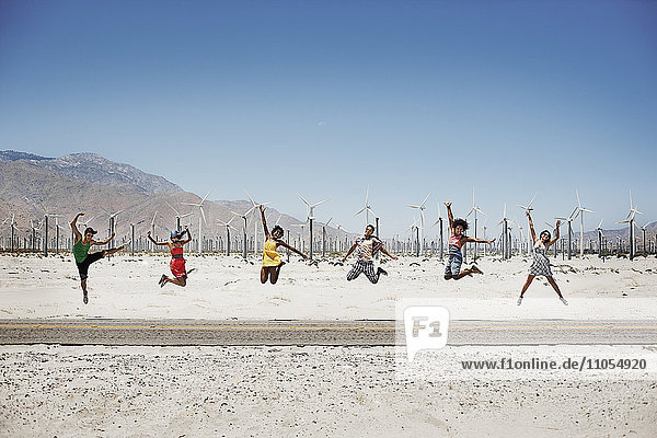 A row of six young people leaping in the air  arms outstretched in wide open space in the desert.