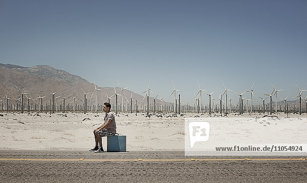 A man standing with a suitcase on the side of a highway  with wind turbines in the background.