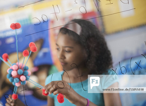A girl holding a molecular structure and looking at a board of equations and formulae in the classroom.