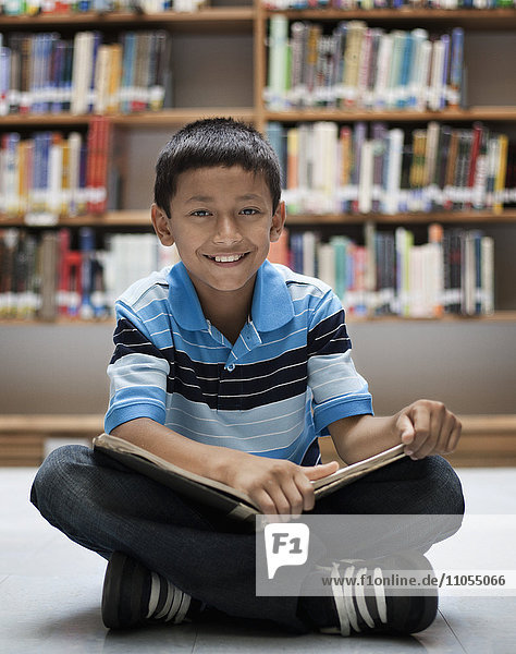 A boy sitting on the floor in a library reading a book.
