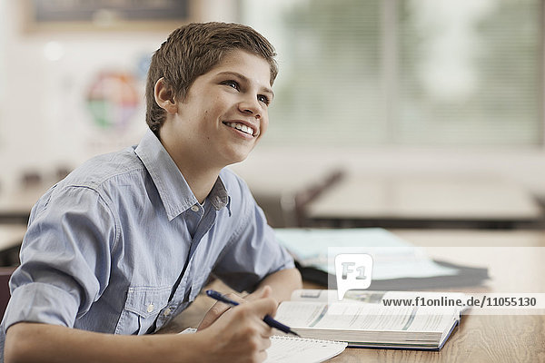 A boy sitting at a desk in class using school books and holding a pen.