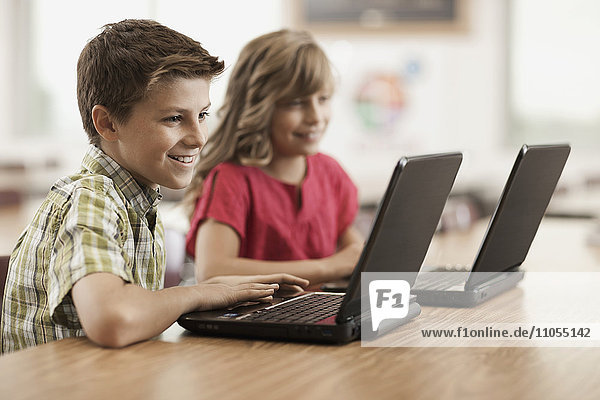 Two children seated at desks in class using laptop computers.