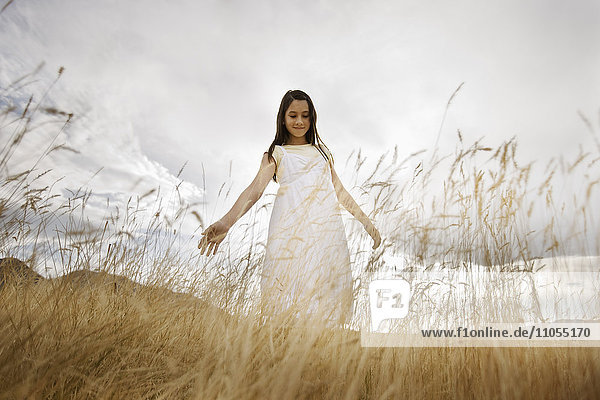 Ein Mädchen in einem weißen Kleid im langen Gras  das mit seitlich ausgestreckten Armen nach unten blickt.