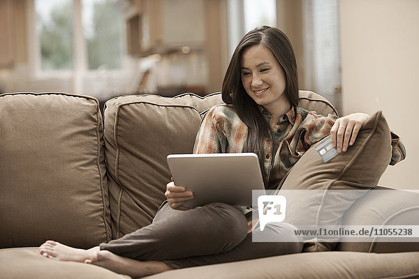 Eine Frau sitzt zu Hause auf einem Sofa  hält ein digitales Tablet und eine Kreditkarte in der Hand und kauft online ein.