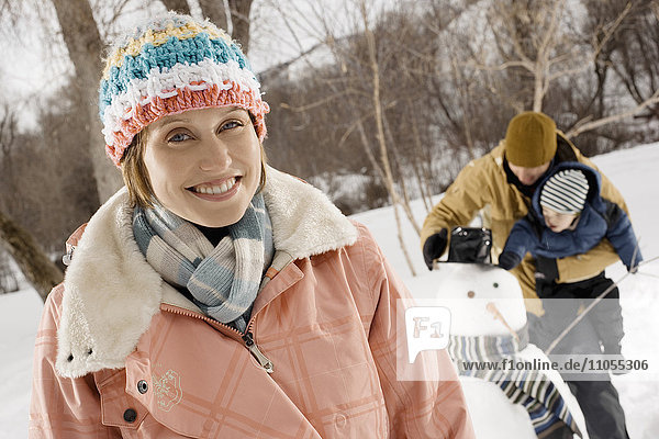 Eine Frau und ein Mann halten ein Kind und inspizieren einen Schneemann mit Hut und Schal.