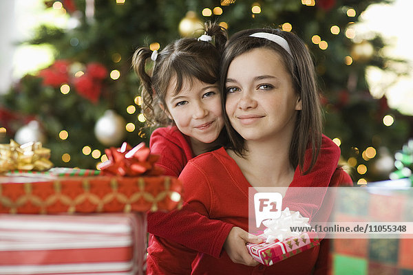 Zwei Mädchen nebeneinander an einem Weihnachtsbaum  umgeben von Geschenken.