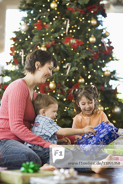 Eine Mutter und zwei Kinder beim Auspacken von Geschenken an einem großen Weihnachtsbaum.