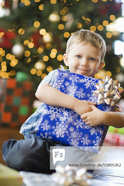 A young boy by a Christmas tree hugging a large wrapped present.