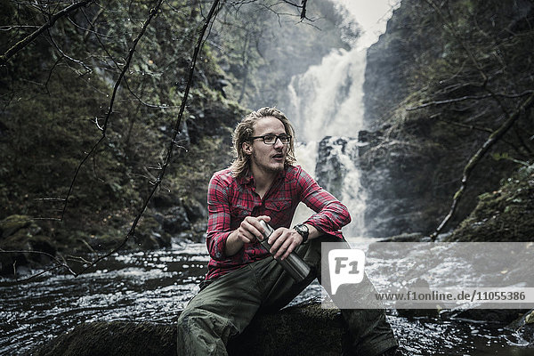 Ein Mann sitzt auf einem Felsen bei einem Wasserfall und hält einen Flachmann. Winterwandern.