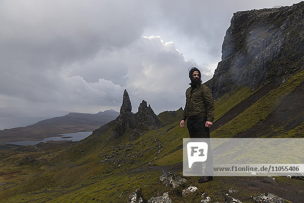 A man on a narrow path rising to a dramatic landscape of rock pinnacles under an overcast sky with low cloud.