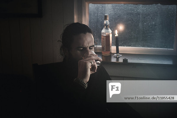 A man sitting in the dark by a window in candlelight drinking from a small glass. A bottle beside him.