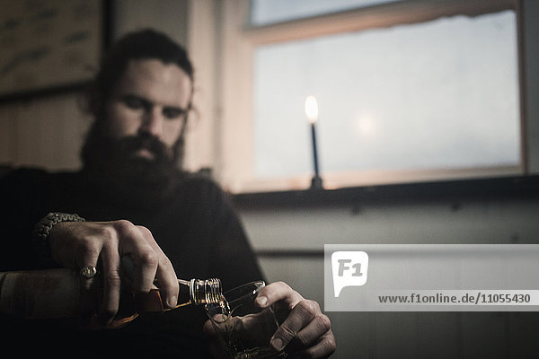 Ein Mann sitzt allein in einem Raum und schenkt sich ein Glas Whisky ein. Eine brennende Kerze.