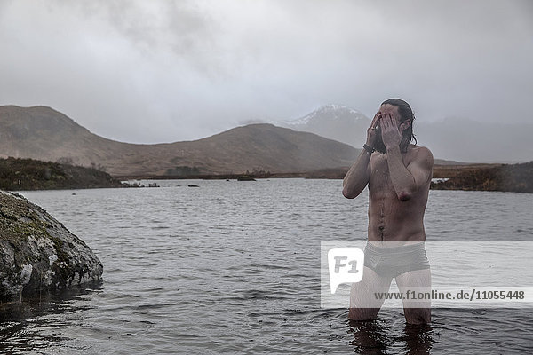 Ein Mann  der mit den Oberschenkeln in Badehosen tief im Meerwasser steht und sich das Wasser aus den Augen wischt.