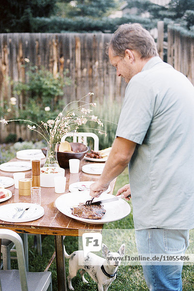 A man carving meat at a family meal in a garden  being watched by a small dog under the table.