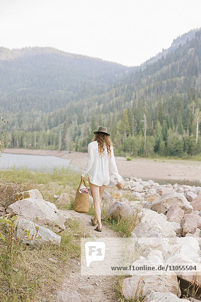 A woman in a hat and white shirt with a basket  by the shore of a mountain lake.